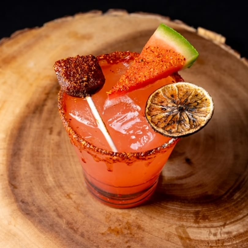 A vibrant red Mexican cocktail with a chili-lime rim, garnished with watermelon, dried lime, and a tamarind candy.