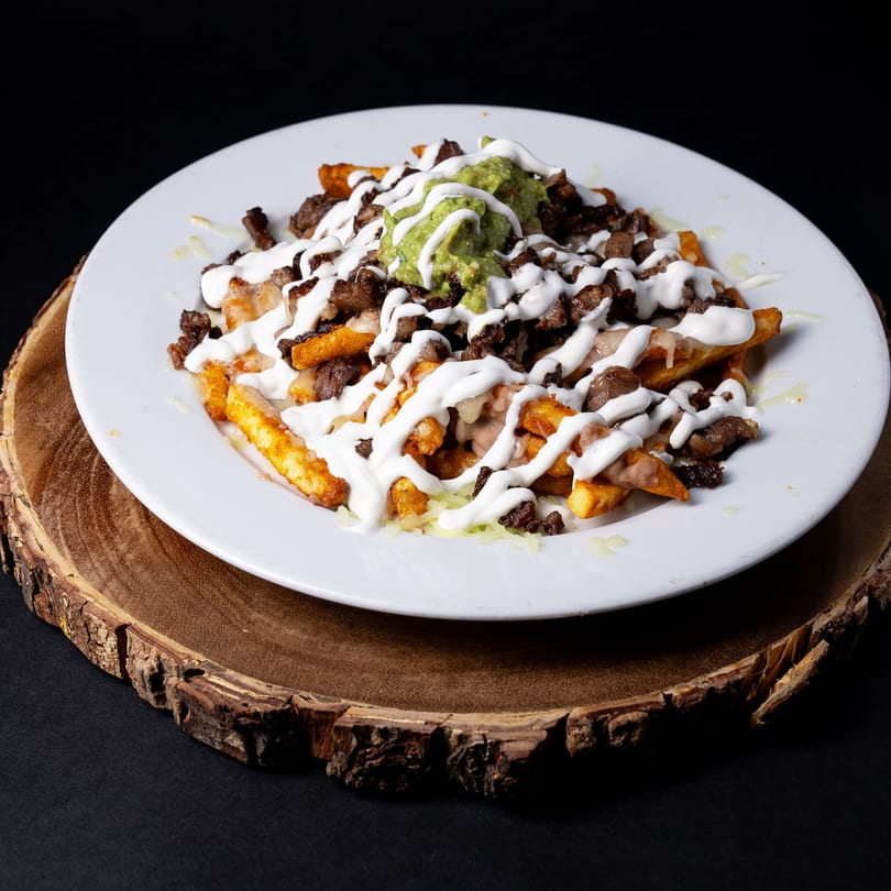 Hecho en Mexico's carne asada fries topped with guacamole, sour cream, and cheese on a white plate.