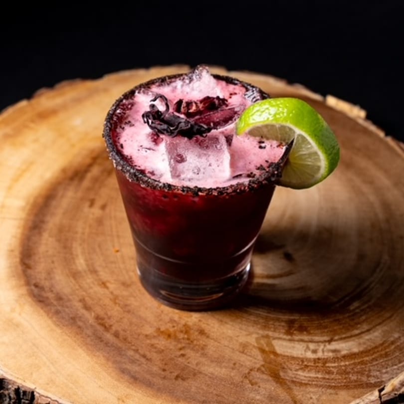 Hecho en Mexico's hibiscus margarita, garnished with a lime wedge and dark rim, on a wooden surface.