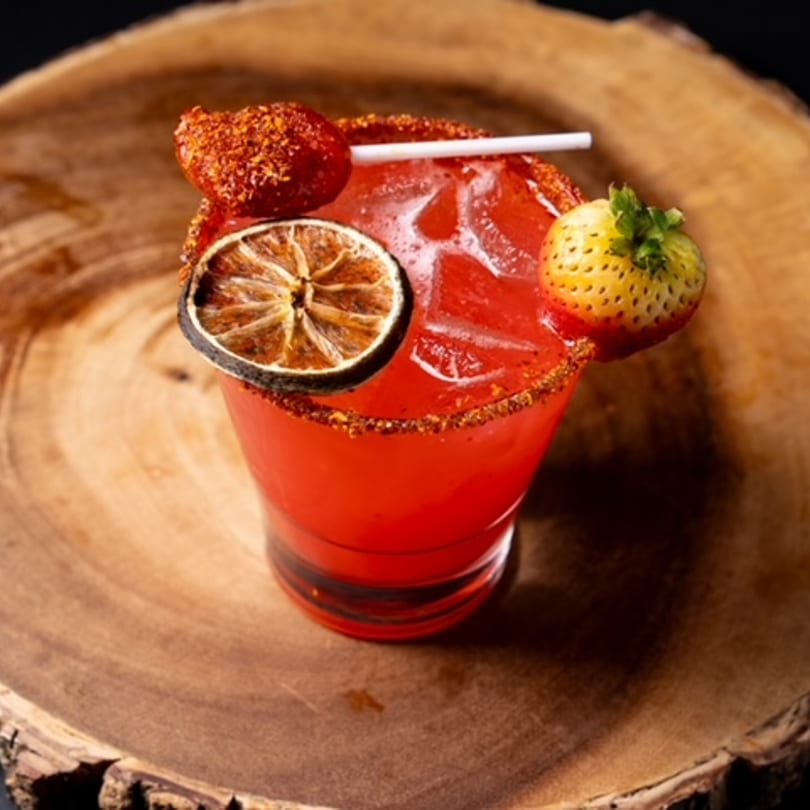 A vibrant red Mexican cocktail with a chili-lime rim, garnished with strawberries and a dried lime slice.