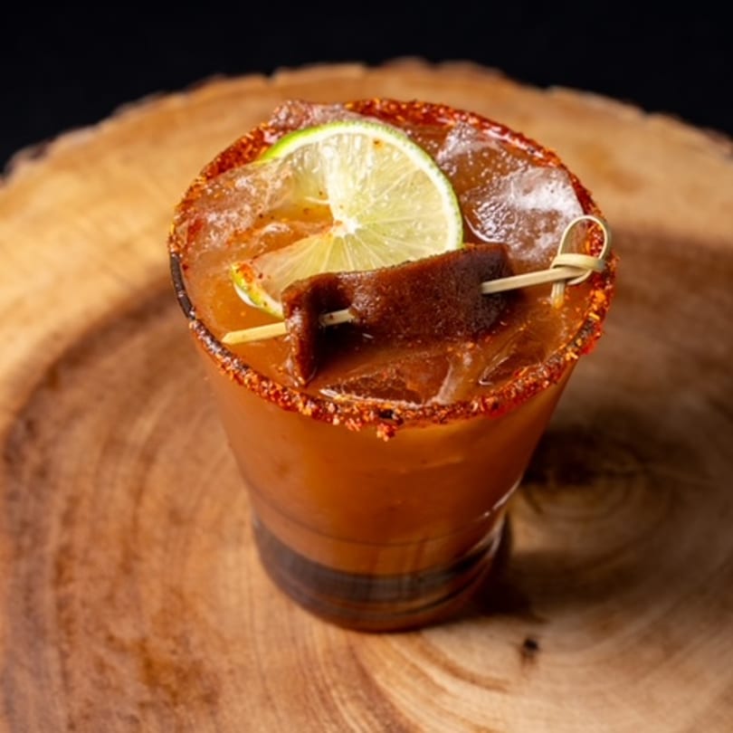 Mexican michelada cocktail with a chili-lime rim, lime slice, and tamarind candy on a wooden surface.