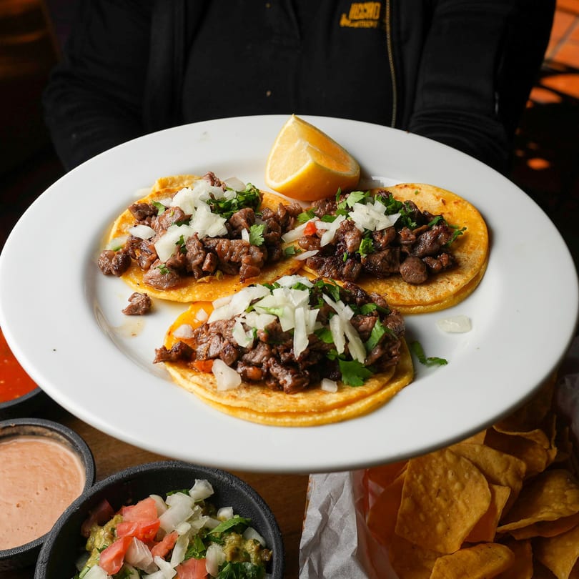 Three carne asada tacos with onions, cilantro, and a lemon wedge on a white plate.