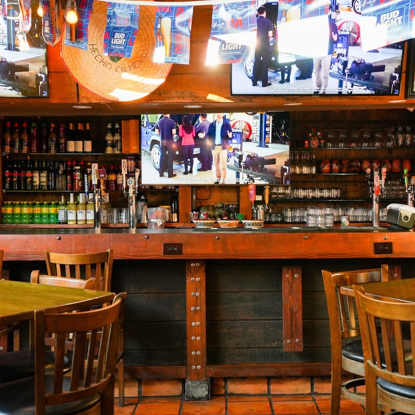 Interior of Hecho en Mexico restaurant with a wooden bar, bottles, beer taps, and TVs showing a car.
