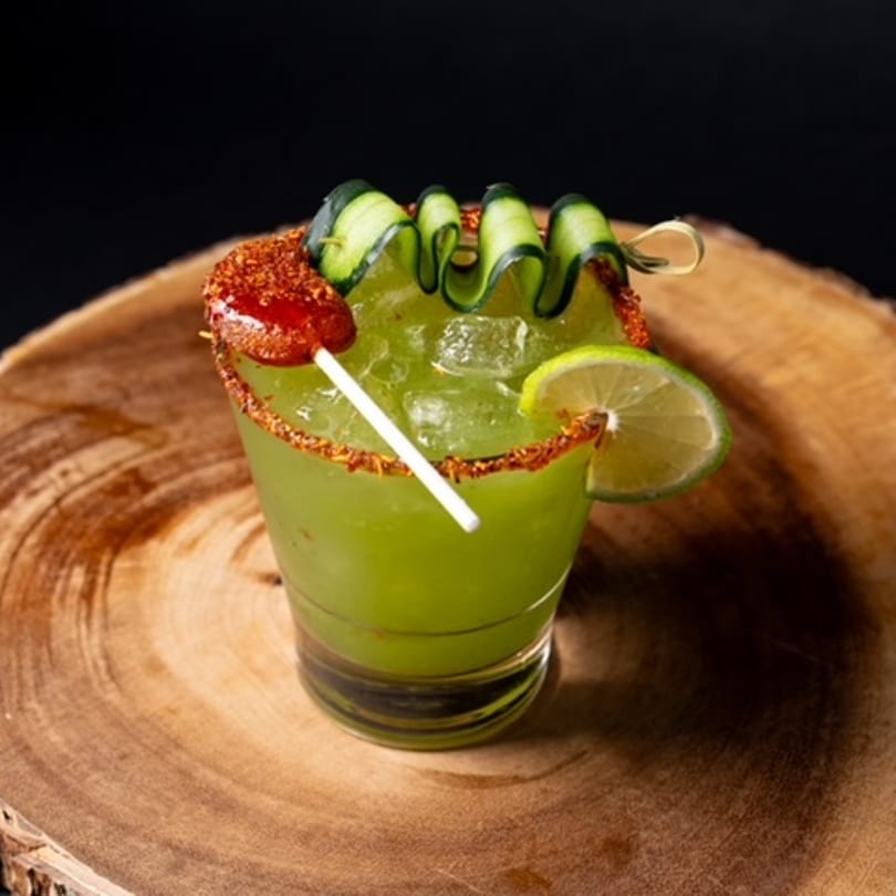 A green Mexican cocktail with cucumber ribbons, chili rim, and a lime slice on a wooden surface.
