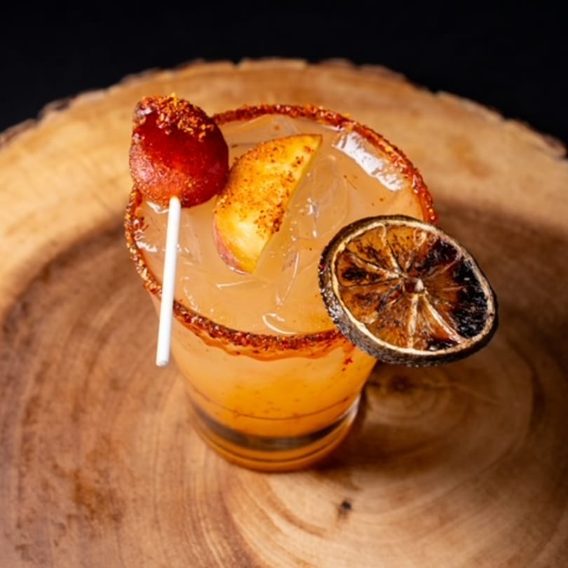 A Mexican cocktail with a chili-lime rim, garnished with a tamarind candy, peach slice, and dried lime.