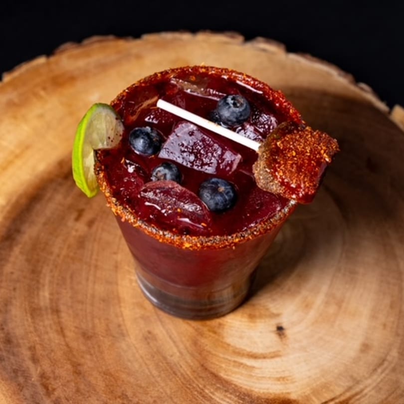 A vibrant red Mexican drink with blueberries, ice, a lime wedge, and a chili-lime rim.