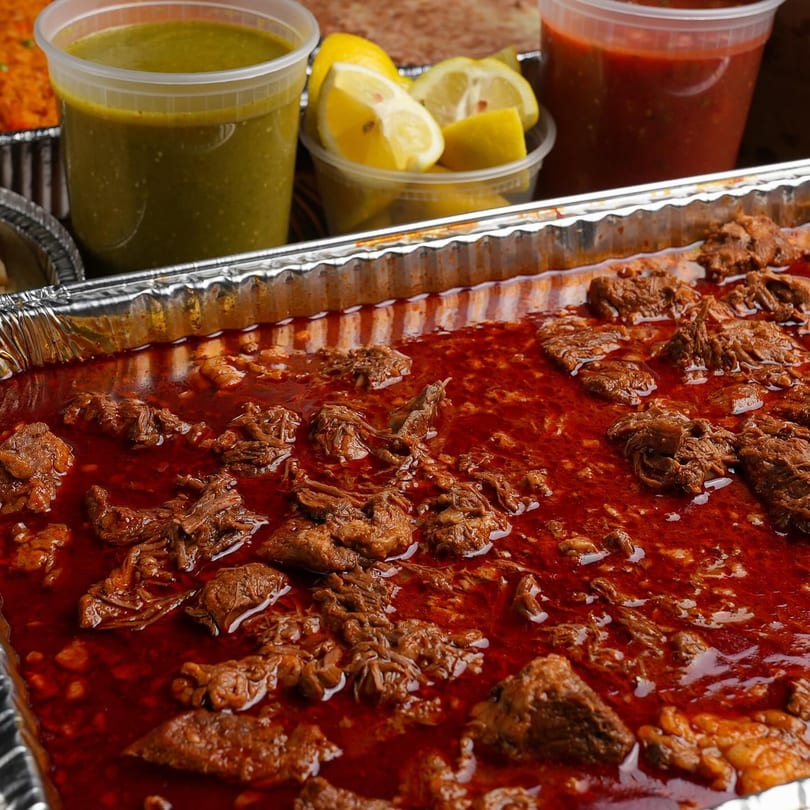 Hecho en Mexico's large catering tray of birria, with sides of green salsa, red salsa, and lemons.