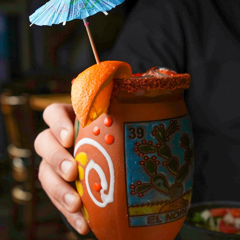A hand holds a traditional Mexican cantarito cocktail garnished with an orange slice and umbrella.