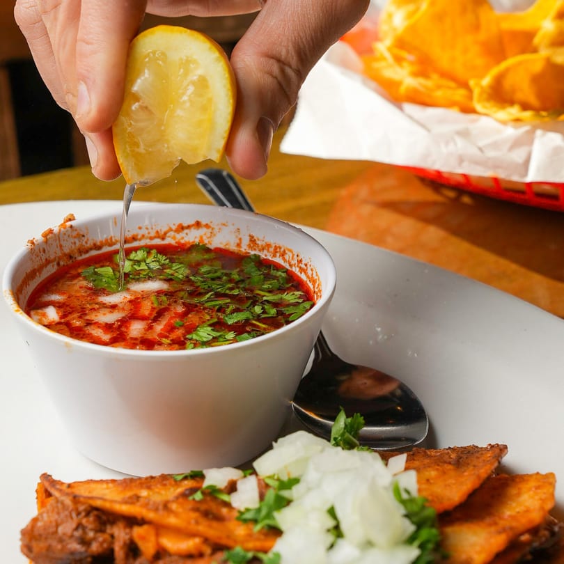 A hand squeezes a lemon over a bowl of birria consommé, with tacos and onions on the side.