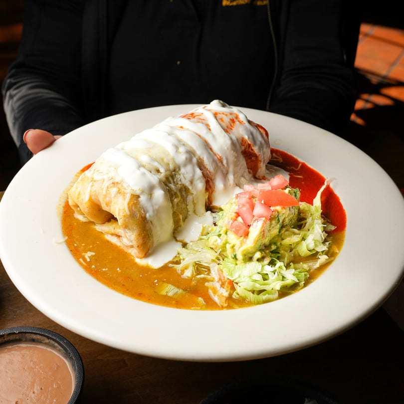 A large, saucy burrito topped with sour cream and red sauce, served with guacamole and lettuce.