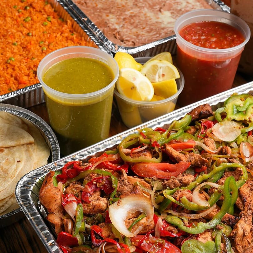 Mexican catering spread with fajitas, rice, beans, tortillas, salsa verde, red salsa, and lemons.