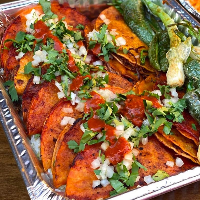 A foil tray filled with birria tacos topped with cilantro, onions, and red sauce, alongside grilled peppers.