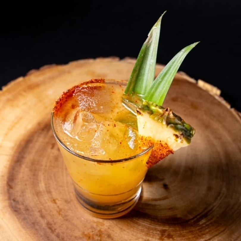 A Mexican pineapple cocktail with a chili-lime rim and pineapple garnish on a wooden surface.