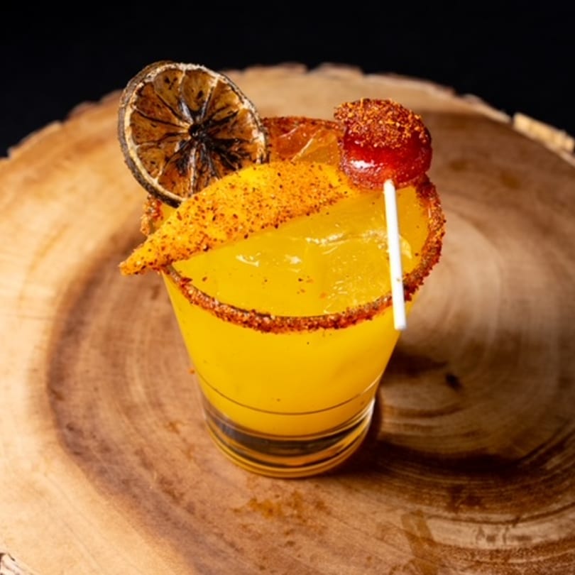 A vibrant yellow Mexican drink, garnished with a dried orange slice, mango, and a chili-dusted lollipop.