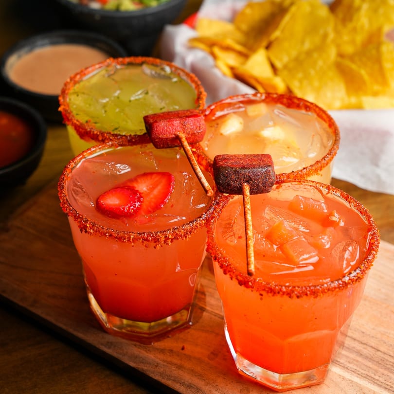 Four vibrant Mexican drinks, two red and two yellow, with chili-rimmed glasses and candy skewers.