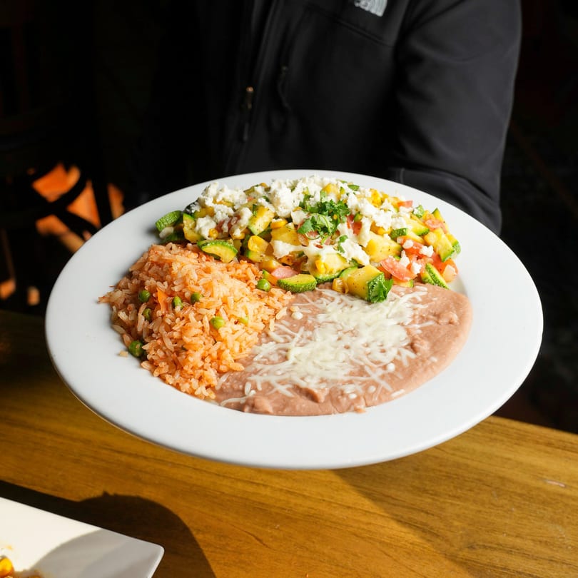 A person holds a white plate with Mexican rice, refried beans, and a vegetable medley.